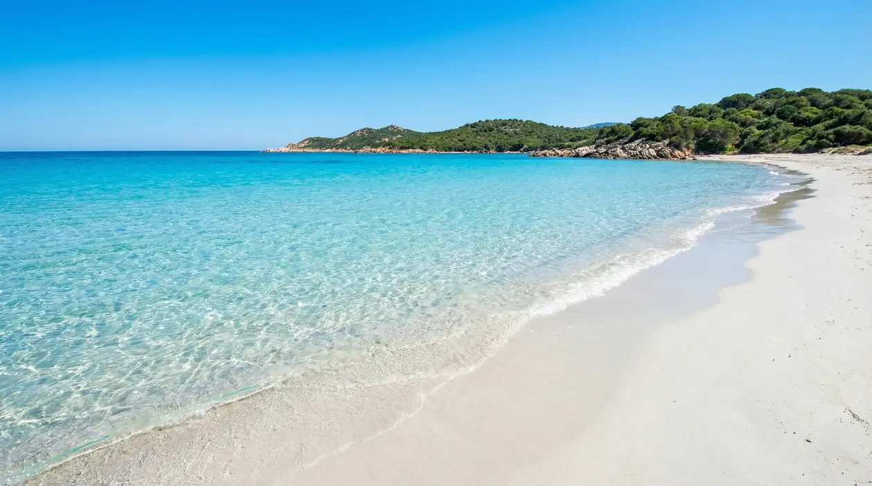 Spiaggia italiana con sabbia chiara, mare cristallino e costa verde in una baia tranquilla
