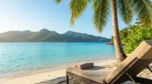 Lettino su una spiaggia tropicale con mare turchese, palme e montagne sullo sfondo