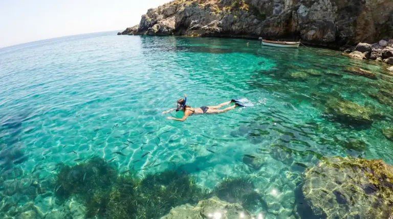 Persona che fa snorkeling in una caletta rocciosa italiana con acqua cristallina e piccoli pesci