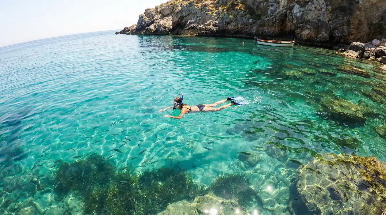 Persona che fa snorkeling in una caletta rocciosa italiana con acqua cristallina e piccoli pesci
